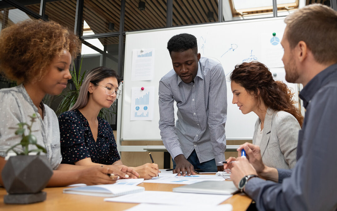 multi-ethic, multi-generational business people gathering around table with whiteboard in background planning